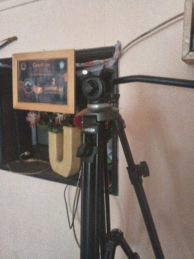 The scene depicts an indoor environment in Dogsenang Gorphoog, Bhutan, focused on a black camera tripod positioned prominently on the right. The tripod features three visible legs, a black pan handle extending to the right, and a camera head with a quick-release plate partially visible, displaying the text "IMAGEN".
Behind the tripod, a dark, horizontal shelf or ledge extends across the background. On this shelf, from left to right, are:
1. A framed certificate with a light wooden frame. The certificate's dark background displays the word "Certificate" at the top and "2 ANNIVERSARY" prominently in the lower section, along with other unreadable text and logos.
2. A small plant with light-colored, possibly artificial, flowers (pink and white) in a dark pot, situated behind a decorative item.
3. A freestanding, light-colored, textured block letter "U".
Several dark cables are visible on and around the shelf, some hanging down behind the tripod. The wall in the background is a light, neutral color, possibly off-white or pale beige, with a visible texture. No individuals are present, and no actions or interactions are depicted beyond the static arrangement of objects. The image quality is somewhat grainy.