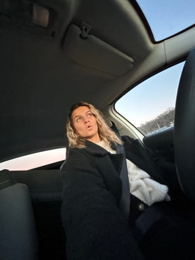 From inside a vehicle, a woman with curly blonde hair appears to be looking upward. She wears a black jacket and a seatbelt, and a white garment is visible. The interior of the car includes a sun visor, a car window, and the dashboard. Outside the car, a bright sky is visible. The perspective is tilted, creating an unconventional viewing angle.