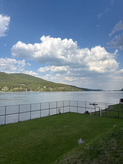 The image shows a body of water, likely a river, with a forested hill on the opposite bank. The sky is blue with large, puffy white clouds. In the foreground, there is a grassy area, possibly a riverbank, with a wire fence. The reflection of the fence is visible on the water. The scene could be in Овсянка, Russia, given the landscape features (river, forested hills) which are consistent with the region's geography.
