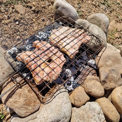 The image shows two pieces of raw chicken on a metal grill over hot coals. The grill is placed on the ground, surrounded by large, smooth river stones. The ground is covered in dry earth, small pebbles, and sparse dry grass. In the background, more dry earth and rocks are visible, suggesting an outdoor, possibly rural or park setting. There is no indication of the city of Gorni Dabnik, Bulgaria. The scene is lit by natural sunlight, casting shadows from the grill and stones.