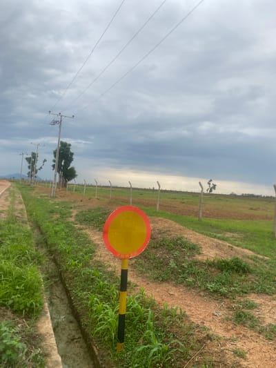The image depicts an outdoor scene in Bulamare, Nigeria, under a cloudy sky, suggesting it is daytime. The perspective is from a low angle, looking upwards and towards a distant horizon. The foreground features a red and yellow circular sign with black stripes on its pole, positioned on the right side of the frame. The ground is a mix of dry soil and patches of green grass.
To the left, power lines stretch across the sky, supported by wooden utility poles. Beyond this, a fence runs along a field that transitions from cultivated patches of brown soil to areas of green vegetation. In the mid-ground, a few scattered trees stand with sparse foliage.
The overall setting appears to be a rural or undeveloped area, possibly on the outskirts of a town or village. The presence of power lines and agricultural fields indicates human activity, though no people are visible in the image. The cloudy weather creates a diffused light, and there is no visible text in the image. The scene evokes a sense of openness and a slightly overcast atmosphere.