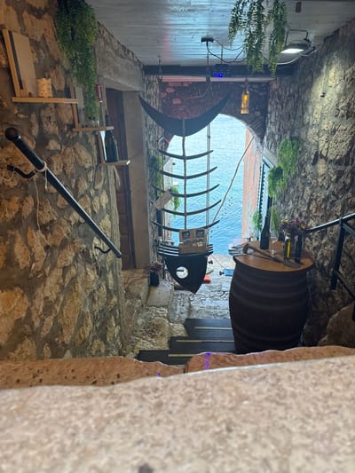 Captured from a close, slightly canted angle, the scene unfolds within a narrow passageway in the city of Grad Rovinj, Croatia. The walls of the alley are predominantly constructed from rough-hewn stone.
In the foreground, a black metal handrail is affixed to the wall. The alley opens up to reveal a view of the sea in the background, framed by the surrounding stone structures. A metal fish skeleton hangs above the passageway, adding a nautical theme to the scene. A wooden barrel, serving as a table, is positioned to the right, adorned with flowers, bottles, and glasses. Green plants are arranged near the top of the passageway. A light fixture illuminates the left side of the alley.