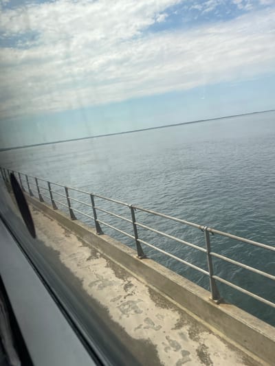 The image captures a view from a train or vehicle moving over a bridge, likely the Great Belt Bridge, in Korsør, Denmark. The foreground shows a section of the bridge's concrete walkway and a metal railing. Beyond the railing, a vast expanse of calm, grey-blue water stretches to the horizon. The sky above is a mix of blue and white clouds, with sunlight filtering through. The overall scene is one of transit over a large body of water.
