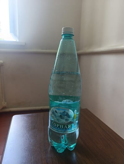 A clear plastic bottle of "Gornaya Prokhlada" mineral water, labeled in Russian, stands on a wooden table. The bottle is partially filled with clear liquid and has water droplets on its exterior. The label features a depiction of mountains and birds, along with text in Russian. The background shows a wall and a window with light filtering through. The scene appears to be indoors, possibly in a room within the city of Gizel, Russia, given the context of the prompt.