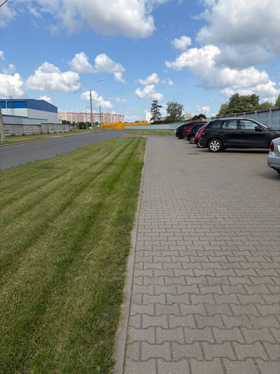 The image captures a view of a parking lot and a roadside area in Navadworski Selski Soviet, Belarus. In the foreground, a paved walkway made of interlocking gray bricks runs parallel to a grassy verge. The grass has distinct mowing patterns, with lighter green stripes indicating recently cut areas. Beyond the grass is an asphalt road.
On the right side of the road, several cars are parked in a line. The closest car visible is a black SUV, followed by a red car, and then other vehicles partially obscured. Beyond the parked cars, a concrete fence runs along the property line, with trees and foliage visible behind it.
In the background, a row of multi-story residential buildings with red brick facades can be seen. To the left, a large building with a blue and white exterior is partially visible. The sky is a bright blue with scattered white, fluffy clouds. Two streetlights are visible along the roadside, one taller than the other. The overall scene depicts an urban or suburban setting on a clear day.