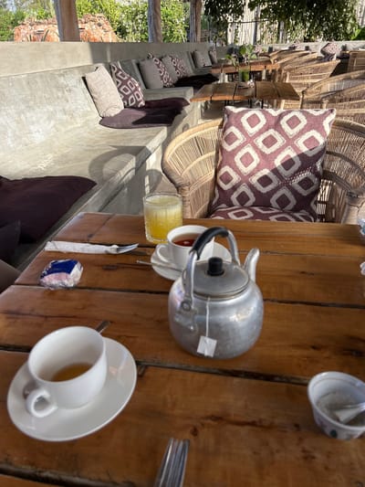 This image depicts a tranquil outdoor cafe or lounge area, likely in Kiwengwa, Tanzania, under bright daylight. The scene showcases a rustic yet comfortable setting, suggesting a place for relaxation and refreshments.
On the right, a substantial wooden table serves as a focal point. It holds a silver metal teapot with a black handle, from which a small white tea bag tag dangles. Nearby, several white ceramic cups are arranged, one containing a dark liquid (likely tea), and another inverted on a saucer. A tall glass filled with a vibrant yellow drink, possibly iced juice or a cocktail, is also present. Further along the table, another white teacup and saucer hold a light-colored beverage. Cutlery, specifically a fork, and a small, blue and white packaged item, perhaps sugar or a napkin, complete the table setting.
To the left, the seating area is composed of tiered concrete benches, generously outfitted with numerous cushions and pillows. Some pillows feature a distinctive brown and white geometric pattern, while others are solid deep maroon or brown, contributing to a cozy ambiance. Beyond these benches, additional seating made of woven natural material, possibly rattan or wicker, is visible, along with more patterned pillows.
The background reveals lush green vegetation and hints of natural fencing or structures, reinforcing the outdoor, open-air environment. The strong, natural light suggests a sunny and clear day, creating an inviting and peaceful atmosphere. No people are visible, and there is no legible text other than the generic tea bag tag and pattern on the small packaged item.
