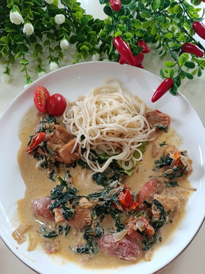 A white plate holds a meal of noodles and a creamy sauce with pieces of meat, spinach, and tomatoes. The noodles are thin and white, piled in the center of the plate. The sauce is a light brown color, and it covers the meat and vegetables, which are arranged around the noodles. Two cherry tomatoes, halved, sit to the left of the noodles, with one whole cherry tomato beside them. In the background, a decorative garland of green leaves and red chili peppers hangs over the plate. The setting appears to be indoors, with a light-colored surface beneath the plate. The lighting suggests it is daytime.