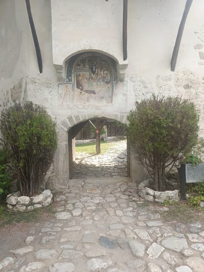 This media file captures an arched stone gateway leading into a courtyard, likely part of a historical religious complex given the fresco above the arch. The fresco depicts a religious scene with figures in traditional attire, rendered in earthy tones. The gateway is flanked by two conical evergreen bushes in stone planters. The pathway leading to and through the arch is paved with irregular cobblestones, suggesting an ancient or traditional construction style. Sunlight streams through the arch, illuminating the courtyard beyond, where a grassy area is visible with some wooden structures and a visible sign. The walls are made of rough stone and plaster, some showing signs of age. The overall impression is one of historical significance and tranquility. The time of day appears to be daytime, with bright sunlight and shadows indicating clear weather. The location is implied to be Rozhen, Bulgaria, consistent with the architectural style and presence of religious iconography. No specific activity or event is depicted, the scene is static and evokes a sense of timelessness. There is a visible sign in the background of the courtyard, but the text is not legible.