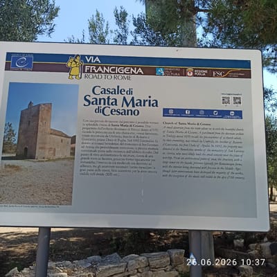 This media file features a signpost with information about the "Via Francigena" and the "Casale di Santa Maria di Cesano." The signpost is placed outdoors, with trees and a clear blue sky visible in the background. In the foreground, there are some stone ruins.
The signpost itself is white with colored sections. It includes text in both Italian and English, detailing the history and significance of the church of Santa Maria di Cesano. The text mentions that the church dates back to 1050 and was rebuilt in 1092. It also describes the architectural style and the frescoes that were once present.
Several logos and emblems are visible on the sign, including those of the "Regione Puglia," "FSC" (Forest Stewardship Council), "Ministero della Cultura," and the "Council of Europe." Social media icons and website addresses are also present, suggesting promotional information for the Via Francigena route. A small, yellow cartoon-like depiction of a pilgrim with a staff and backpack is featured on the left side of the sign.
The image also includes a timestamp in the bottom right corner: "26.06.2026 10:37". This suggests the image was taken on June 26, 2026, at 10:37 AM. The lighting indicates it is daytime, likely morning. The overall scene conveys a historical and cultural context, highlighting a specific point of interest along the Via Francigena pilgrimage route.