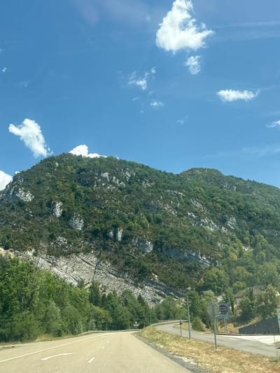 This image depicts a scenic view of a mountain range and a road in the vicinity of Villard-Saint-Sauveur, France. The mountain is covered in lush green vegetation, with exposed rocky strata visible on its slopes. Several white clouds dot the clear blue sky. A paved road with white lane markings curves through the foreground, suggesting a driving perspective. On the right side of the road, there are streetlights, road signs, and some buildings nestled amongst the trees. The overall impression is one of a natural landscape encountered while traveling.