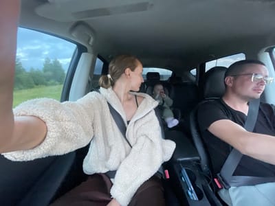 A family of three is traveling in a car on a cloudy day. The driver, a man wearing glasses and a black t-shirt, is seen from the side. Next to him, a woman with her hair pulled back in a bun, wearing a cream-colored fleece jacket over a black top, is looking back towards the rear of the car. In the back seat, a baby is secured in a car seat, facing forward. The car appears to be on a road in a rural area, with trees and a grassy field visible outside the windows. The interior of the car shows a car console with cupholders and a charging cable. The sky is overcast, suggesting an impending storm or a generally gloomy weather condition. The location is specified as Мещеряки, Russia.