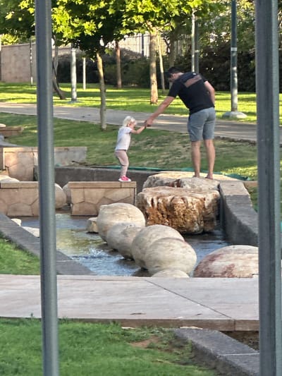 In the city of Givat Shmuel, Israel, a father and his young daughter are interacting in an outdoor park with a water feature. The father, wearing a dark t-shirt and light blue shorts, is standing on a large, rough-textured rock. He is reaching out his right hand to his daughter, who is standing on a lower, flat-topped stone. The daughter, with blonde hair and wearing a light-colored shirt and pink leggings, is reaching out her left hand to grasp her father's. The water feature consists of smooth, rounded stones submerged in shallow water, with larger stone structures forming the edges of the area. Green grass and trees surround the water feature, and a paved pathway is visible in the background. Two tall, grey poles, possibly part of a lighting or infrastructure system, are in the foreground, partially obscuring the view. The scene appears to be during daylight, with soft, natural light illuminating the area.