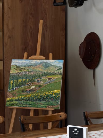 The image depicts an indoor scene with a painting displayed on an easel. The painting is a landscape with rolling hills, a village, trees, and a field of sunflowers in the foreground. The painting is signed and dated "Dsegh 2023". To the right of the easel, a brown cowboy hat is hanging on a pale wall. In the foreground, a wooden table is visible, with a portion of a napkin holder featuring a silhouette of a man. The overall setting appears to be a domestic or artistic space. There are no people visible in the image. The scene is static and devoid of action.