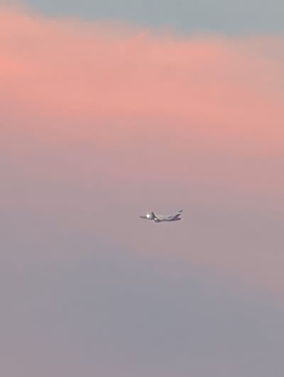 An airplane is captured in flight against a soft, pastel sky. The sky transitions from a pale blue at the top to a warm pink, suggesting either sunrise or sunset. The airplane, which appears to be a commercial jet, is seen from a side profile and is positioned in the lower half of the frame, slightly to the left of center. It is a small detail in the vast expanse of the sky. The image does not depict any specific landmarks or elements that would place it in the city of Böblingen, Germany. The environment is solely the sky.