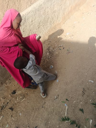 This image depicts two individuals outdoors in Gobirawa, Nigeria, near a light-colored, textured earthen wall. An adult female, partially visible, is seated or crouched, wearing a vibrant red or magenta full-body garment and a patterned blue fabric beneath. Her right arm shows a light-colored wrist accessory. A young child, wearing a light grey tunic and trousers with light-colored open-toed footwear, stands leaning towards the female and holds onto her red garment with their right hand. The female is looking downwards and to her left, towards the child or an unseen point. The ground is exposed light brown soil or sand, covered with scattered debris including paper scraps, plastic fragments, and organic matter. Shadows of individuals are visible on the ground, indicating direct sunlight.