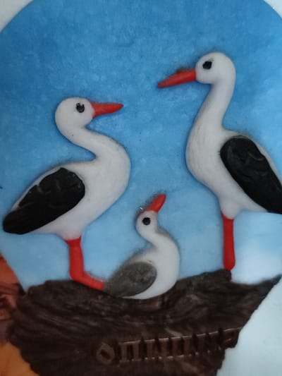 The image shows a close-up of a decorative object featuring three storks. Two adult storks, white with black wings and red legs and beaks, are positioned on the right and left of the frame, facing inward towards each other. Between them, a smaller, juvenile stork, also white with gray coloring on its back and neck, faces towards the left adult stork. The storks are perched on a dark brown, textured surface that appears to be a stylized representation of a nest or ground. The background is a solid, textured light blue. The object is likely a figurine or a relief. There is no visible indication of the city of Voskobiinyky, Ukraine within the image itself.