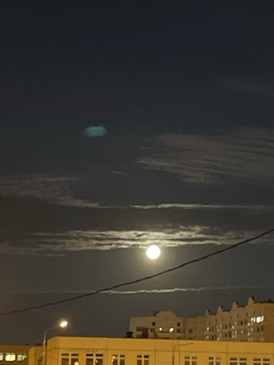 The image captures a nighttime scene in the city of Zakharino, Russia. The sky is dark, with the moon prominently visible, partially obscured by wispy clouds. A faint, greenish rectangular object is also present in the upper portion of the sky. Below the sky, buildings with illuminated windows and streetlights are visible, suggesting an urban environment. A power line cuts diagonally across the frame. No people are discernible in the image.