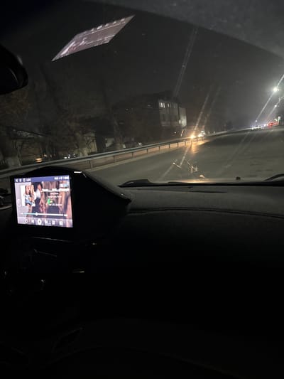A nighttime view from inside a car, showing a highway through the windshield. Headlights and streetlights illuminate the road. The dashboard includes a screen displaying images or video. The scene is mostly dark, emphasizing the contrast between the illuminated highway and the interior.