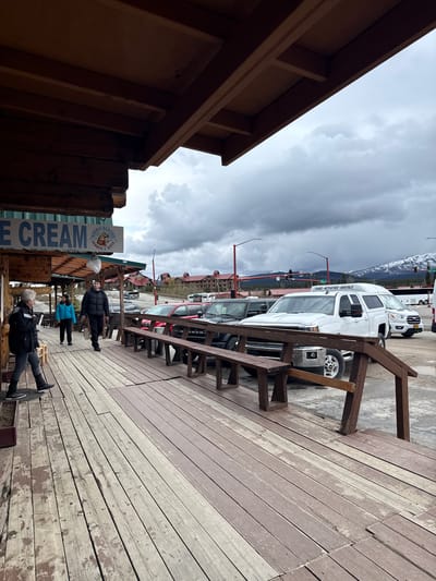 The image shows an outdoor scene on a cloudy day in Healy, United States. A weathered wooden deck stretches into the foreground, with people walking or standing on it. An "ICE CREAM" sign is visible on a wooden structure. Cars and vans are parked along the street, with buildings and distant mountains visible in the background under a sky filled with heavy, dark clouds.