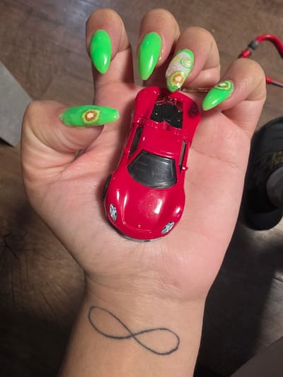 A close-up shot, taken in Bremerhaven, Germany, features a person's right hand holding a small, glossy red toy sports car. The hand is positioned palm-up, with the bright red car resting centrally on the palm, oriented with its front towards the wrist.

The fingernails are long, with a distinct pointed (stiletto or almond) shape, and are painted a vibrant neon green. Several nails are adorned with fruit-themed decals: the middle finger nail has a white base featuring multiple slices of various fruits, including kiwi, lime, and lemon. The pinky finger nail and the index finger nail each display a prominent kiwi slice decal, while the thumb and ring finger nails are a solid neon green.

On the inner side of the wrist, a simple black outline tattoo is visible, resembling an infinity symbol or a stylized number eight. The background is blurred and dark, suggesting an indoor setting, with muted tones of brown that could be wood and a visible, out-of-focus red object or strap in the upper left corner. No text is visible in the image.