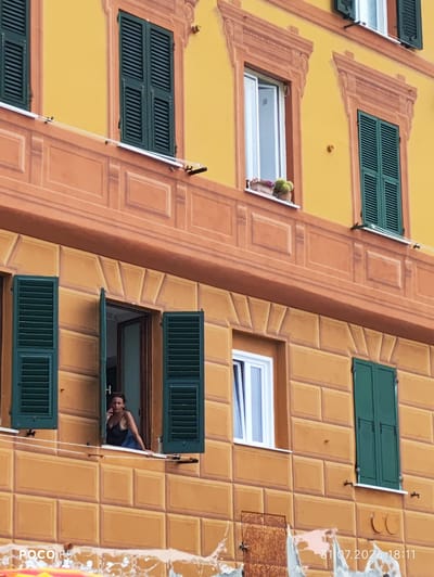 A woman in a black tank top is leaning out of an open window, talking on a cell phone. She is positioned on the second floor of a building with yellow and orange painted walls, featuring decorative stucco work and green shutters. There are multiple windows visible on the facade, some open and some closed. The building appears to be in a Mediterranean coastal town, likely Camogli, Italy, given the architectural style and vibrant colors.