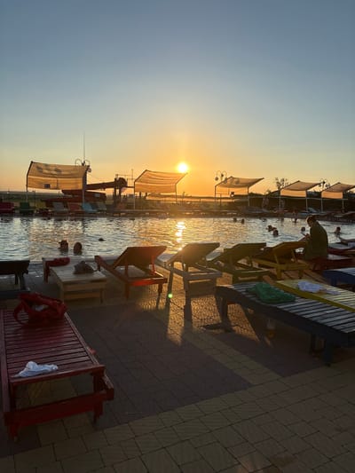 The image depicts an outdoor swimming pool area with people enjoying the water. The sun is setting, casting a warm golden light across the scene and reflecting off the rippling water. Several individuals are visible in the pool, some swimming and others relaxing. Along the edge of the pool, there are lounge chairs and tables, with some people seated or standing on the wooden deck. Two lounge chairs, one red and one yellow, are prominently placed near the foreground. Red towels or clothing are draped over some of the furniture. The setting appears to be a leisure or resort area, with awnings providing shade over parts of the seating area. The ground is paved with interlocking bricks. The time of day is clearly evening, indicated by the low angle of the sun and the warm, diffused light. The weather appears to be clear and pleasant. There is no visible text in the image. The overall mood of the image is one of relaxation and enjoyment on a summer evening.