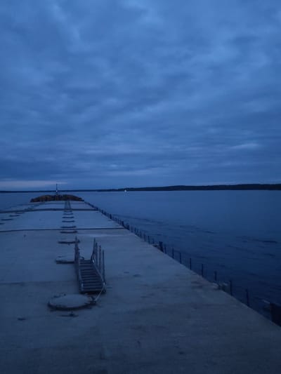 The image depicts a concrete pier extending into a body of water under a cloudy, twilight sky. The pier has a series of rectangular segments separated by seams, as well as metal access covers. A set of metal stairs leads down to a lower level. A line of vertical posts runs along the pier's edge. The opposite shore is visible in the distance. The location is indeterminate.