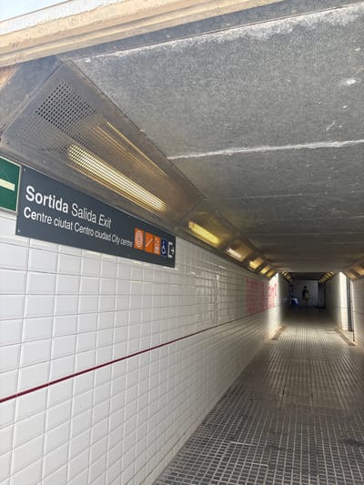 The image shows an underground pedestrian walkway in Vilassar de Mar, Spain. The walkway is lined with white tiled walls and a grey tiled floor. A sign above the left wall reads "Sortida Salida Exit" and displays various icons, including those for accessibility, fire safety, and a wheelchair. There are fluorescent lights illuminating the walkway. In the distance, two figures are visible, one appearing to be a woman with a bag. The overall impression is of a functional, utilitarian public space.