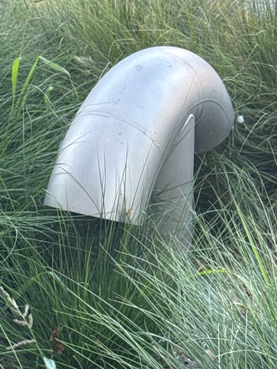 A light gray, curved pipe structure emerges from tall, dry grass in Strombeek-Bever, Belgium. The pipe has a smooth, metallic surface with visible seams. The surrounding environment is densely covered with thin, wispy blades of grass, some of which are light green and others appear dry and tan. There are no people or animals visible in the image.