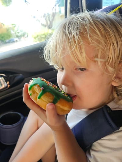 A child with blonde hair, secured in a car seat with a black shoulder strap, is captured in an eye-level, close-up shot as they lick a donut with green icing and multi-colored sprinkles. The child's face is positioned towards the donut, obscuring most of their features except for their eye, nose, and the tip of their tongue. The background includes a portion of the car's interior, including a cup holder and a blurred view of the outside environment. The image suggests the scene is taking place in a vehicle, potentially in transit.