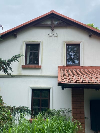 The image shows the exterior of a white house in Albertowsko, Poland. The house has a reddish-brown tiled roof and several windows with dark frames. One of the windows is partially covered by a decorative shutter with a wavy pattern. The numbers "1910" are visible in white on the wall of the house, indicating its construction year. Green foliage, including tall grasses and evergreen bushes, surrounds the house. The sky appears overcast, suggesting cloudy weather. The scene is a still shot, capturing the architecture and surrounding vegetation of a residential property. There is no activity or people visible in the image.