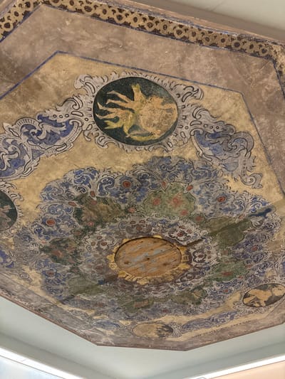 The image depicts a detailed view of an ornate, multi-panel painted ceiling within the Papadopoulos Mansion (Konstantinidis Mansion) in Drama, Greece. The ceiling features an octagonal central field surrounded by trapezoidal panels, all adorned with intricate artwork.
The central part of the ceiling displays a prominent, circular wooden boss or medallion, severely aged and worn, revealing its natural wood grain and concentric marks. Surrounding this, and extending across the panels, is a complex decorative scheme dominated by flowing, stylized floral and vegetative motifs, primarily in shades of muted blue, gray, green, and red, set against a golden-beige background. These patterns include acanthus leaves and various stylized blossoms.
Several circular medallions are integrated into the design. One at the top-center of the frame prominently features a stylized, possibly mythological, yellowish-green figure or creature with multiple limbs or tendrils, set against a dark background. Other similar, less distinct medallions are visible towards the lower edges. The perimeter of the ceiling is framed by a darker, repeating geometric border, possibly a chain-link or looped pattern.
The artwork exhibits significant signs of age and degradation, including discoloration, fading, visible cracks in the plaster, and areas where paint has flaked off, particularly around the central wooden element and some of the medallions. The overall impression is one of historical preservation with evident wear. A modern, plain white ceiling or wall section is visible along the bottom edge of the primary image, indicating the surrounding architecture or a recent intervention. No people are present in the scene.