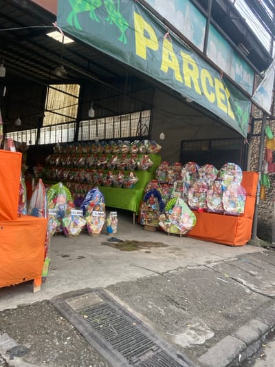 The image shows a storefront with a sign that says "PARCEL" in large yellow letters on a green banner with cartoon frogs. The storefront is located in Bojongsoang, Indonesia (latitude: -6.971, longitude: 107.637). The storefront is a semi-open space with a concrete floor. The storefront is selling gift baskets, which are arranged on shelves and in rows. The baskets are filled with various items, including food and other goods. Some of the baskets have price tags attached to them. There is an orange tarp covering the front of the store. There is a drain in the foreground. The image was taken on a clear day, with no people visible.