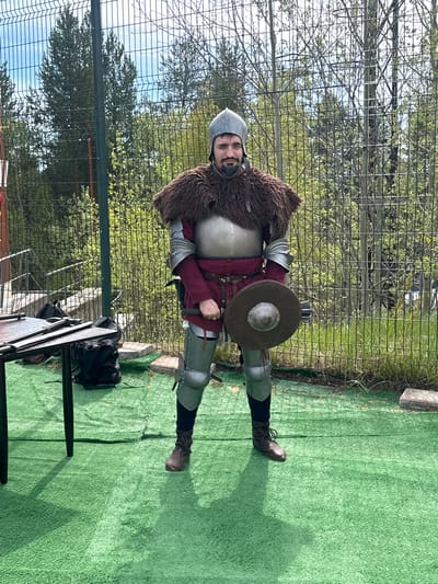 A man dressed in medieval armor stands on a green artificial turf surface. The armor includes a helmet, breastplate, gauntlets, and leg protection. He is also wearing a fur collar and a burgundy tunic. He holds a wooden shield in his left hand. A chain-link fence is directly behind him, with trees visible beyond. To the left of the man, a table with equipment is visible. The photograph was taken during the day, with bright sunlight. The city of Polarnie Zori, Russia cannot be confirmed as the location.