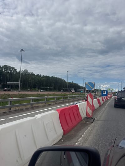 The image captures a view from inside a vehicle looking out onto a highway. In the foreground, the side mirror and a portion of the car's interior are visible. The highway is bordered by white and red plastic barriers, suggesting ongoing road work or a temporary traffic control setup. Several cars are present on the highway, moving in the same direction as the viewer.

Further down the highway, there's a blue structure with a white framed window, possibly a small temporary office or control booth. A large blue road sign with a white arrow pointing left and down is mounted on a stand. In the distance, a dense line of trees forms the background, with tall lampposts lining the highway. The sky is overcast with gray clouds. The location is identified as Levashovo, Russia.