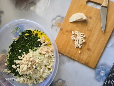 In a top-down view of a kitchen scene, a clear plastic bowl overflows with chopped ingredients, including green herbs, diced yellow bell peppers, and white onions, interspersed with small cubes of what appears to be cheese or tofu. To the right, a wooden cutting board displays several slices of the same white cubed ingredient and a chef's knife with a silver blade and a light-colored wooden handle. The background consists of a light-colored, possibly tablecloth surface with a floral pattern. A subtle watermark at the bottom left corner reads "Galaxy S24 Ultra". The scene is brightly lit, suggesting it was captured indoors. There is no indication of the city of Tilekmat, Kyrgyzstan in the image.