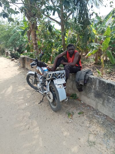 A black male adult, wearing a red tank top and dark pants, sits on a concrete wall. He is looking directly at the camera. A silver motorcycle, with the license plate number "188 LNXX 7530", is parked next to him. The motorcycle is facing away from the camera. A grey jacket lies on the wall near the man. The scene is a dirt road, with a concrete wall bordering the road. The image appears to have been taken in a rural part of Sapele, Nigeria. The location is approximately 5.86 degrees latitude and 5.701 degrees longitude. There are trees and plants in the background.