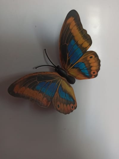 An artificial butterfly with orange and blue wings is positioned on a light-colored, smooth surface. The butterfly's body is black. The wings display intricate patterns, with dark veins on the orange sections and a series of blue patches. Two black antennae extend from the head. The lighting suggests the object is indoors, with a shadow cast to the right. There is no indication of the city of Promyshlennaya, Russia.