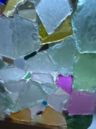 The image shows a close-up of a mosaic made of irregular pieces of colored glass, possibly sea glass, fused together with a clear material. The glass pieces vary in size, shape, and color, including shades of white, blue, green, yellow, and pink. The mosaic has a textured, three-dimensional appearance due to the varied thickness and arrangement of the glass fragments. There are no discernible people, actions, or specific environmental elements like a city or geographic location present in this abstract close-up. Therefore, information about Ridabu, Norway is not applicable to this image.