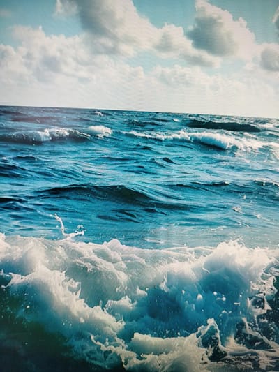 A close-up, eye-level shot captures a dynamic ocean scene under a partly cloudy sky. The water is a deep, rich blue, with white foam cresting on the waves, particularly in the foreground where a large wave is breaking. The sky above is a lighter blue, with scattered, fluffy white clouds. There are no people or man-made objects visible in the image, and no identifiable landmarks to suggest a specific location within Yashkino, Russia. The focus is entirely on the natural elements of the sea and sky.