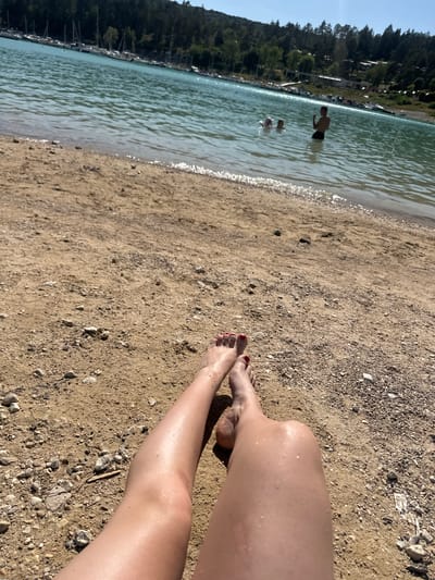 The image shows a person's legs and feet resting on a sandy beach next to a body of water. The water is a clear turquoise color and appears to be a lake, with a line of boats docked in the distance. Trees and a hillside form the background. In the water, three people are visible, two of whom appear to be children or adolescents, and one adult male. The sun is shining, and the overall scene suggests a leisurely day at the beach. The location is identified as the town of Maisod, France.