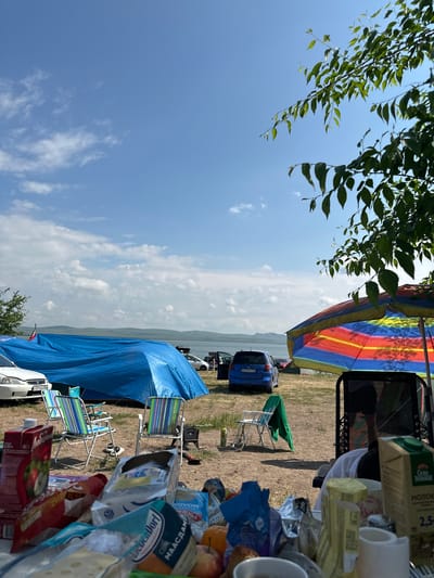 The image depicts an outdoor scene, likely a picnic or campsite, under a partly cloudy blue sky. In the foreground, a table is laden with various food items and beverages, including a carton of juice, bags of snacks, and a package of cheese. Several camping chairs are set up, one with a colorful umbrella providing shade. A large blue tarp covers what appears to be a tent or shelter, with several vehicles parked nearby, including a white car and a blue minivan. In the background, a body of water is visible, with hills or mountains in the distance. The environment appears to be a grassy area, possibly near a lake or river. There are no visible people in the image, and the scene suggests a relaxed, recreational atmosphere. The location is identified as Kolodezny, Russia.
