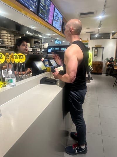 In a brightly lit indoor establishment, possibly a restaurant or food service counter, a bald man with muscular arms is performing a push-up. He is positioned horizontally, with his hands on a light-colored countertop and his body extended. He wears a dark tank top and dark pants, along with black athletic shoes with red laces.
Behind him, the scene appears to be a service counter. A digital menu board displays text and images, including the phrase "MAKE IT A MEAL". Another sign reads "Auténtico Mexicano Delicioso". Several employees are visible behind the counter, interacting with customers or working with equipment. One employee, wearing a black cap and a yellow garment, looks up from behind the counter. There are also digital screens and what appears to be a cash register or payment terminal. In the background, another person in a neon yellow shirt is also seen, and further back, two people are seated, one on a stool. The overall impression is a busy, casual food service environment. There is a sign indicating "SALSA STATION" twice in the background. The context provided indicates the location is North Boambee Valley, Australia, suggesting this is a local eatery. The time of day is not explicitly evident, but the lighting suggests it's indoors during operating hours.