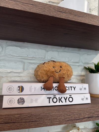 A brown, fuzzy plush toy with two brown legs sticks out from between two white books. The plush toy has a smiling face with two black dots for eyes and a curved black line for a mouth. The books are placed vertically on a wooden shelf. The spine of the left book reads "MICCITY2" in large black letters. The spine of the right book reads "TOKYO" in large black letters. Both books have smaller text and circular emblems on their spines. The background appears to be a white brick wall with some greenery visible at the top. The lighting suggests daytime. The overall scene is a close-up of a shelf with decorative items.