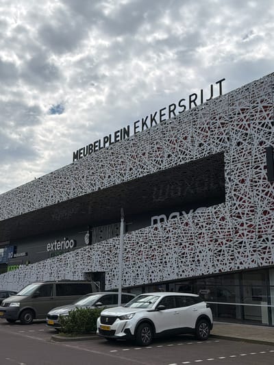 The image captures the exterior of a large retail building with the prominent signage "MEUBELPLEIN EKKERSRIJT" across its upper facade. The building features a distinctive white geometric patterned screen covering much of its surface, with dark horizontal bands and large glass windows at ground level. Several vehicles are parked in front of the building, including a dark grey van on the left, a silver sedan in the middle, and a white SUV on the right. The context provided identifies the location as Son en Breugel, Netherlands, specifically within the Ekkersrijt retail area which is known for furniture stores. The sky is overcast with scattered clouds. The scene appears to be an outdoor daytime shot of a commercial complex.