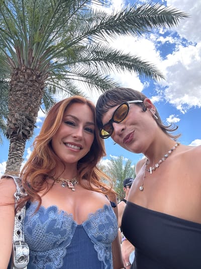 Two individuals pose for a selfie outdoors in Coachella, California, USA. A red-haired woman smiles at the camera, wearing a blue lace top and a silver belt. Above her, another person wears dark sunglasses, a black top, and a pearl necklace. A palm tree is to the left of the frame, with a blue sky and clouds visible in the background.