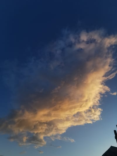 This media file shows a dramatic cloud formation against a deep blue sky, likely at sunset. The clouds are illuminated with warm, golden light on their edges, creating a striking contrast with the darker, shadowed portions and the surrounding blue sky. Some smaller, wispy clouds are visible lower in the frame, and a silhouetted structure, possibly a building or utility pole, is visible on the right edge of the image. The overall scene evokes a sense of natural beauty and the awe-inspiring power of the sky. The time of day is likely late afternoon or early evening, given the lighting on the clouds. There is no visible text in the image.