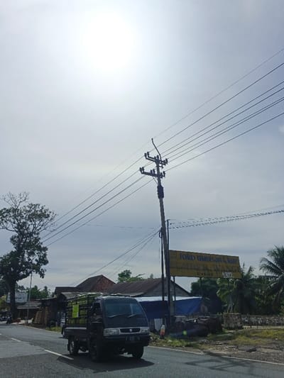 A daylight street scene shows a black pickup truck driving on a paved road under a bright, overcast sky. The sun, high in the sky, is surrounded by a faint solar halo.
In the foreground, a black, older model pickup truck with a white Suzuki emblem on its grille moves from left to right. Its cargo bed is loaded with several stacks of green cylindrical objects, possibly gas cylinders or large bottles. The truck's license plate, partially visible, reads "F 9092 YA."
To the right of the road stands a tall wooden utility pole with multiple cross arms holding numerous electrical wires that stretch across the frame. Below the power pole, a faded yellow sign is mounted on a separate pole. Visible text on the sign includes "TOKO MANISAN & K*re" (with part of 'Kare' obscured), "DIANJUR," and "1319." "Toko Manisan" is an Indonesian term for a sweet shop or grocery store, and "Dianjur" is a regency in West Java, Indonesia.
In the background, behind the sign and pole, are several modest buildings with red-tiled roofs, partially obscured by lush green foliage, including palm trees on the far right. To the far left, a large deciduous tree with sparse leaves stands prominently. The overall setting appears to be a rural or semi-urban area. No people are clearly visible in the image.
