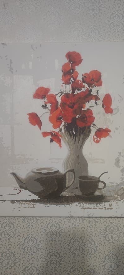 The image displays a painting of a still life featuring a bouquet of red poppies in a vase. To the left of the vase, there is a teapot and a teacup with a spoon. The background is a light, textured gray, possibly simulating a wall. The foreground shows a surface with a patterned wallpaper. The painting is hung on a wall which also has a patterned wallpaper, in a style consistent with interior decoration. There are no people or animals visible in the painting or in the surrounding environment. The scene depicts a domestic setting, likely a room. There is no information available about the location of this image in the city of Svitlovodsk, Ukraine.