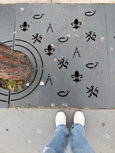 The image shows a close-up, high-angle view of a patterned metal grate surrounding a tree trunk, as seen from a pedestrian's perspective. The grate, a dark gray color, features cut-out patterns of fleur-de-lis, compasses, and smiling lips. The tree trunk, on the left side of the frame, is rough and brown, with some green foliage at its base. The grate is set into a concrete sidewalk. The lower portion of the image shows the feet of an individual wearing blue jeans and white sneakers, standing on the concrete. The overall scene appears to be an outdoor urban environment, possibly in Roermond, Netherlands, given the context of the request. The lighting is natural and the image is clear, with no overt signs of activity beyond the implied presence of the photographer.